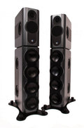 A tall, slim floor-standing Kii THREE BXT active loudspeaker column in a dark grey finish, formed by a compact Kii THREE monitor mounted on top of the taller BXT bass-extension module.