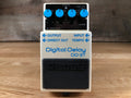 White digital delay pedal with blue knobs and labeled input/output jacks on a wooden surface.