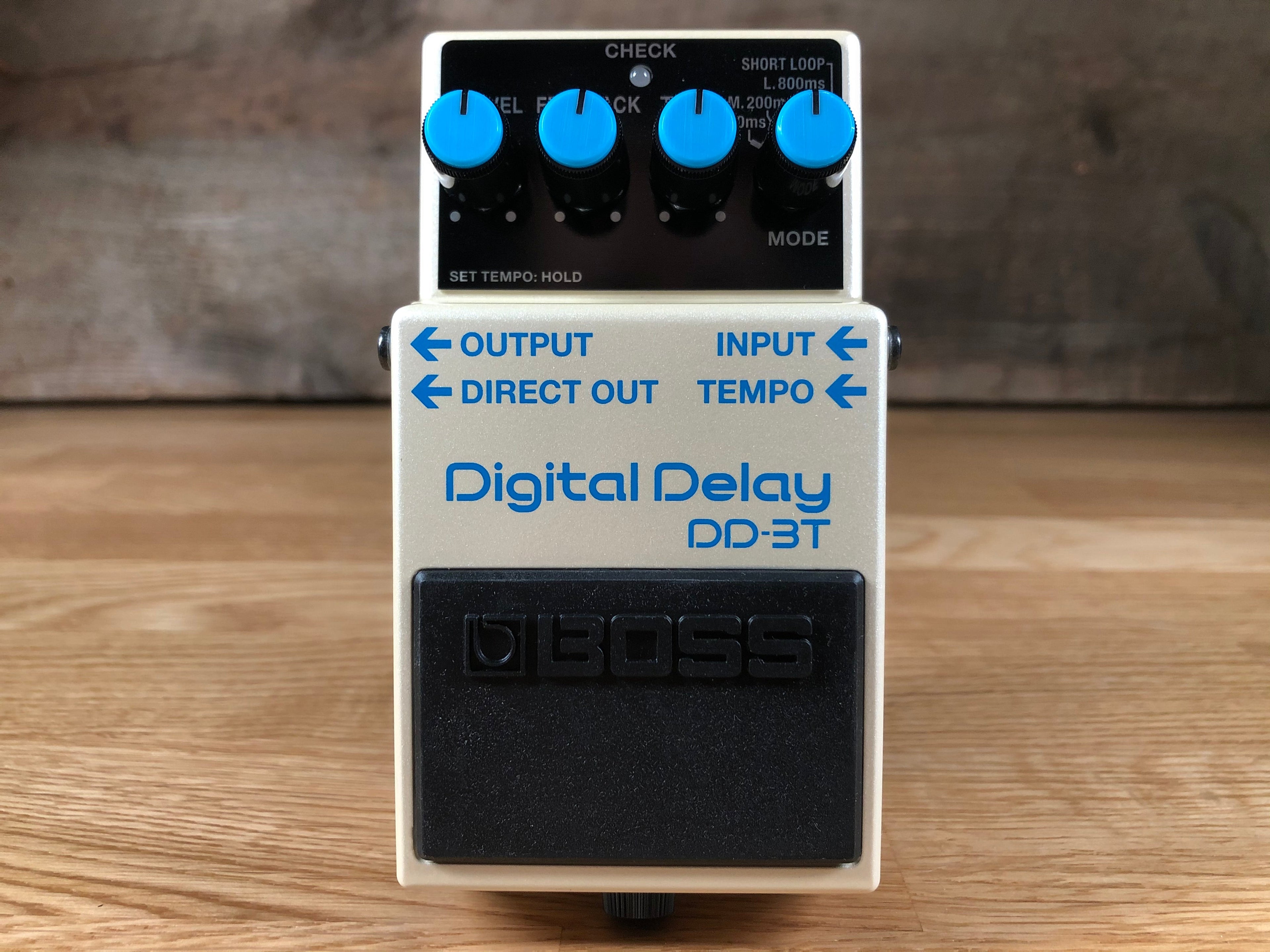 White digital delay pedal with blue knobs and labeled input/output jacks on a wooden surface.
