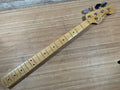Maple guitar neck with metal frets and tuning pegs labeled Fender Precision Bass resting on wooden surface.