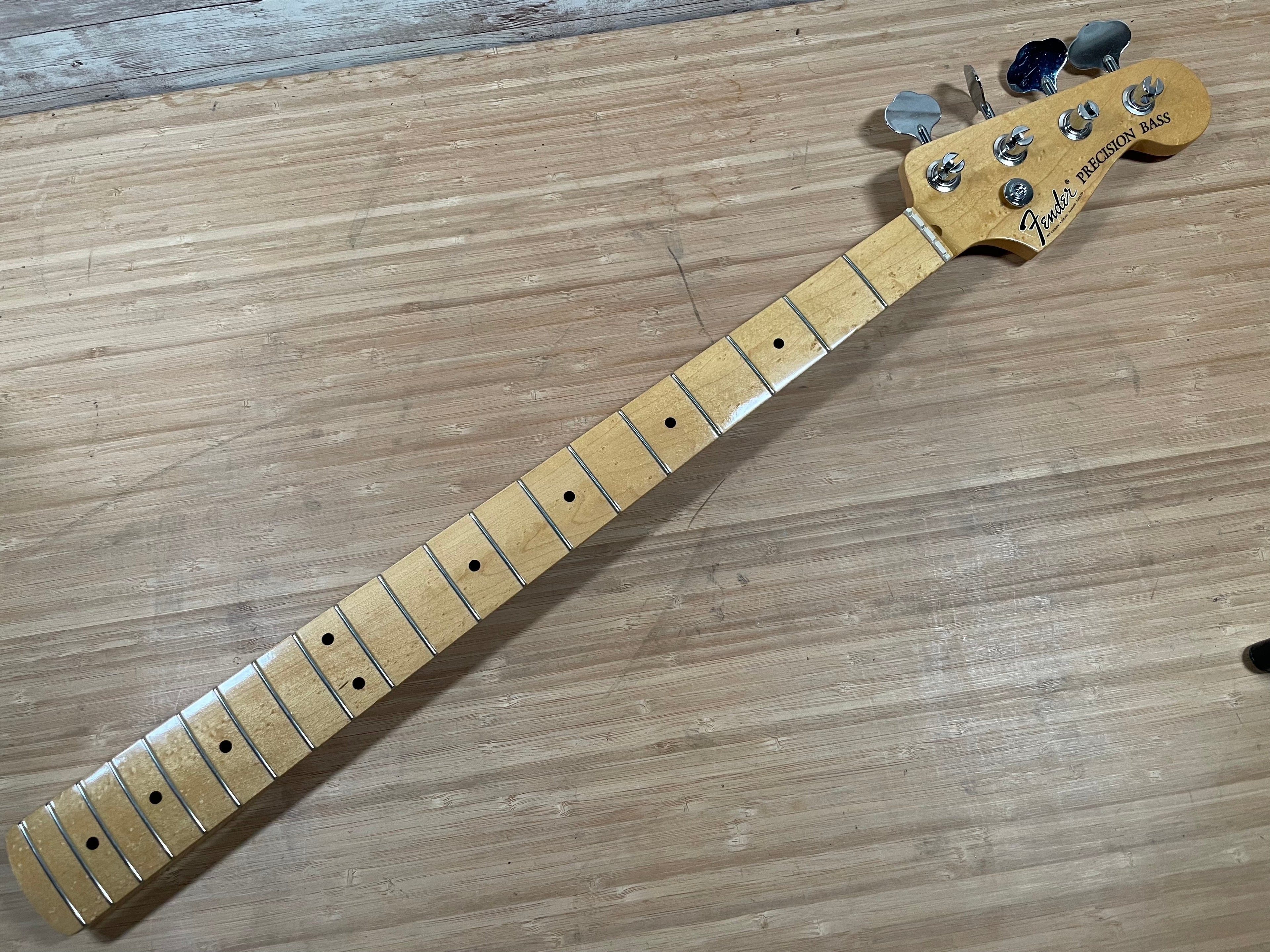 Maple guitar neck with metal frets and tuning pegs labeled Fender Precision Bass resting on wooden surface.