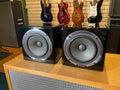 A pair of black cube-shaped Avantone studio monitor speakers placed on a wooden surface with guitars in the background.