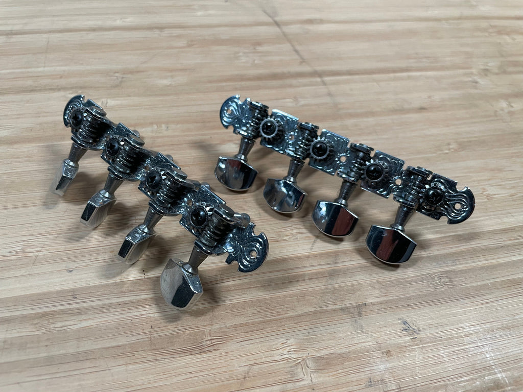 Two sets of four silver metal plate tuners with black screws lay on a wooden surface.