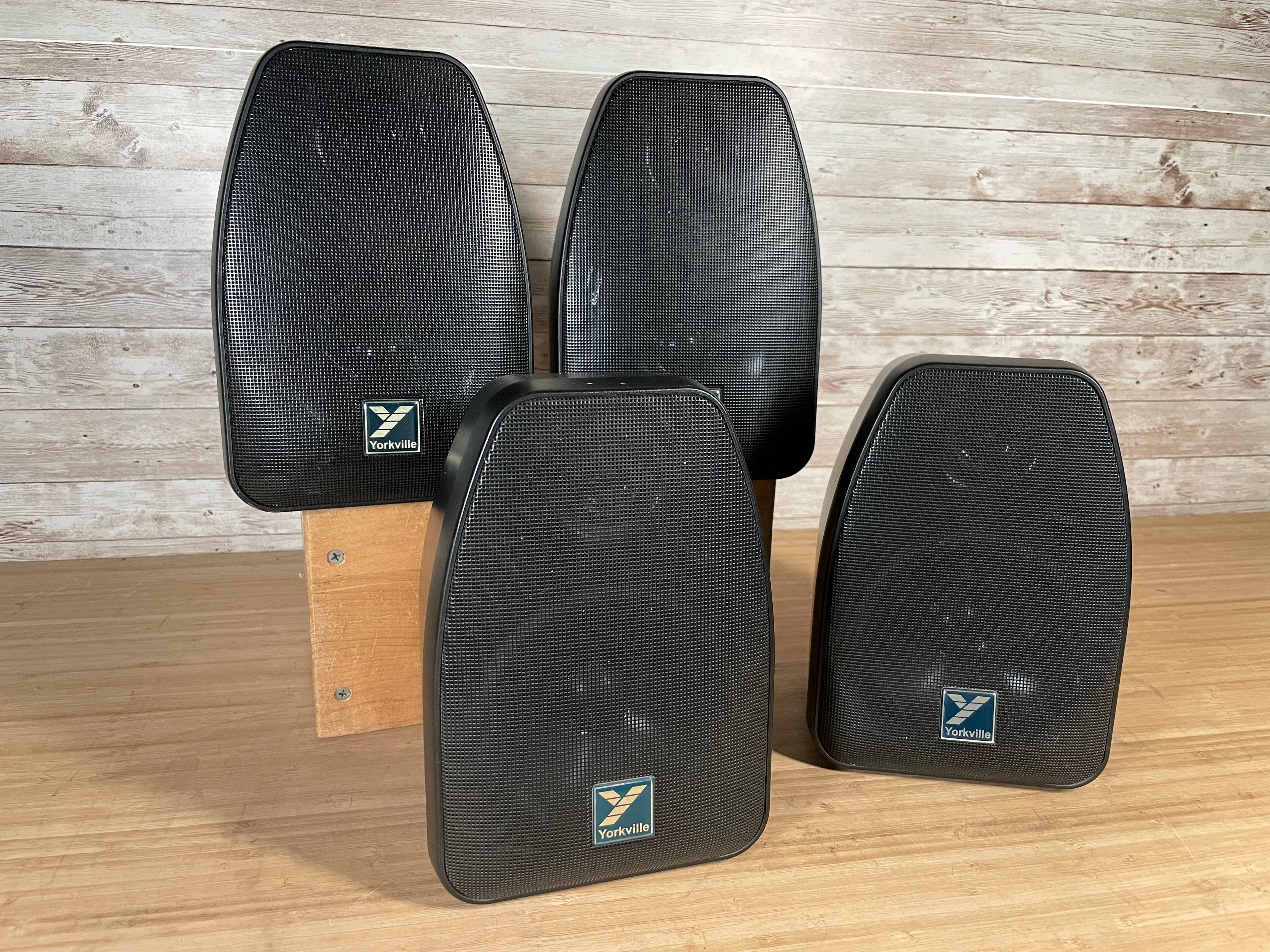 Four black Yorkville speakers with front mesh grills arranged on a wooden surface and wood stand.