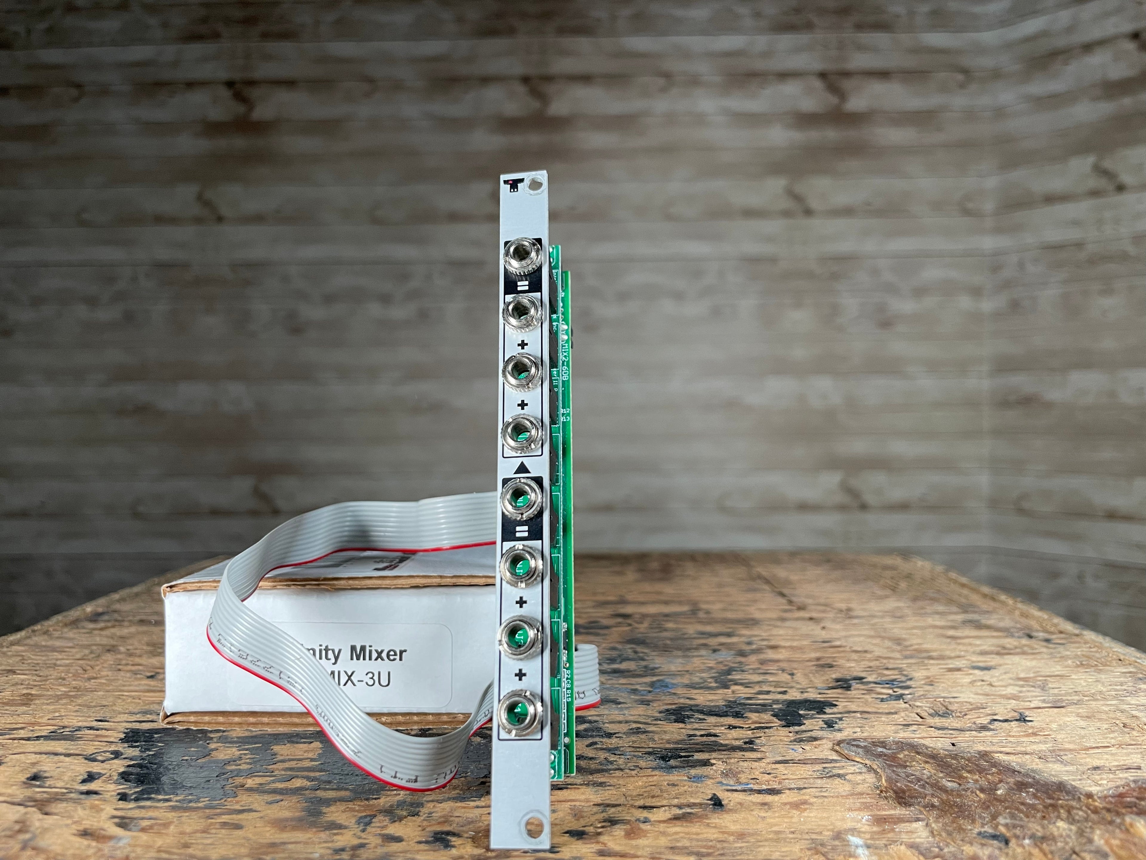 Eurorack module with six jack sockets and a ribbon cable connected, standing next to a cardboard box labeled Unity Mixer MIX-3U.