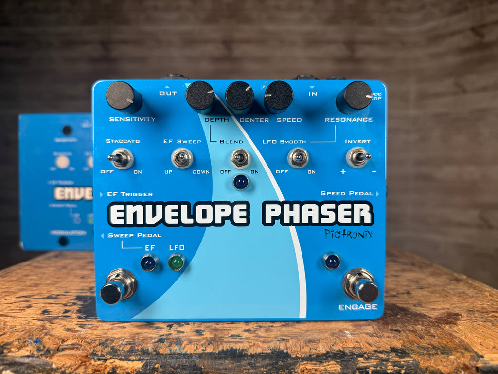 Blue Pigtronix Envelope Phaser pedal with knobs, switches, footswitches, and original box behind it on wooden surface.