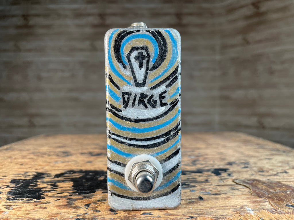 Small rectangular metal device with a toggle switch and artistic painted design with the word DIRGE.