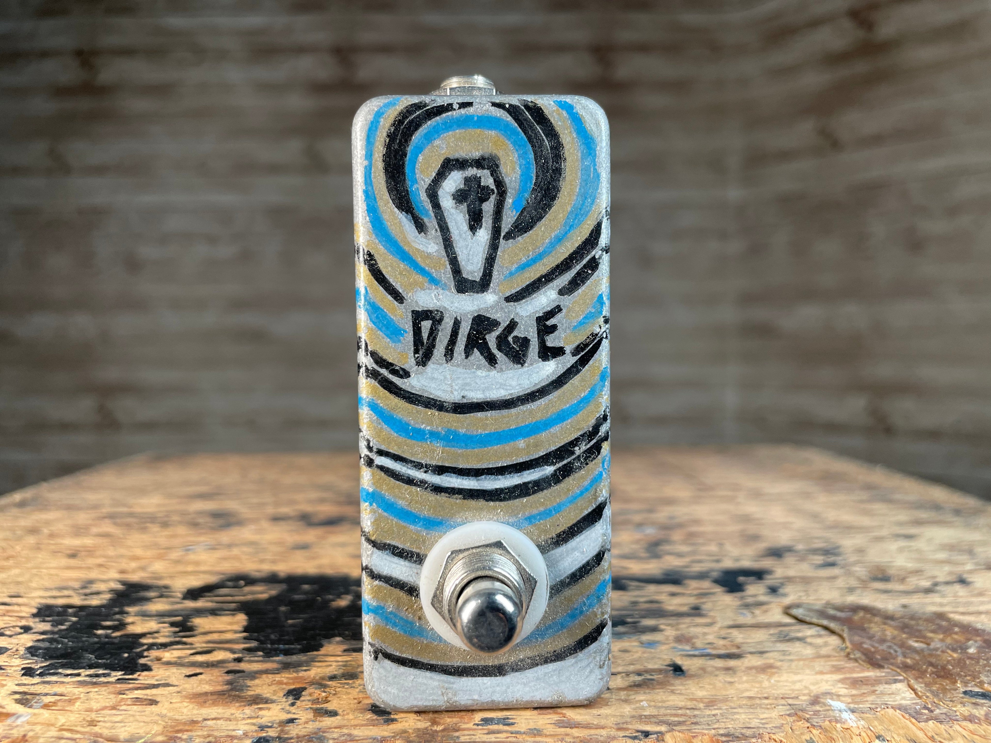 Small rectangular metal device with a toggle switch and artistic painted design with the word DIRGE.