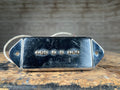 Metal guitar pickup with dog-ear mounting ears and attached white wire, shown on a wooden surface.