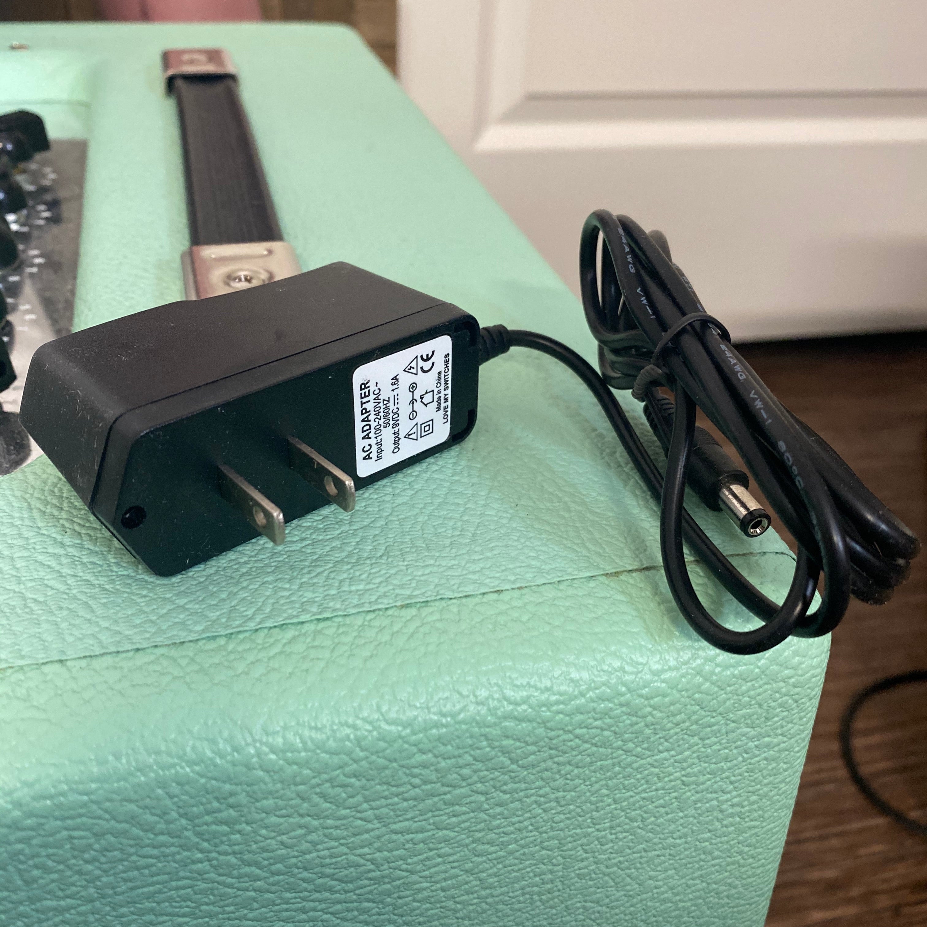 Black AC power adapter with US plug and attached cable with barrel connector on aqua textured surface.