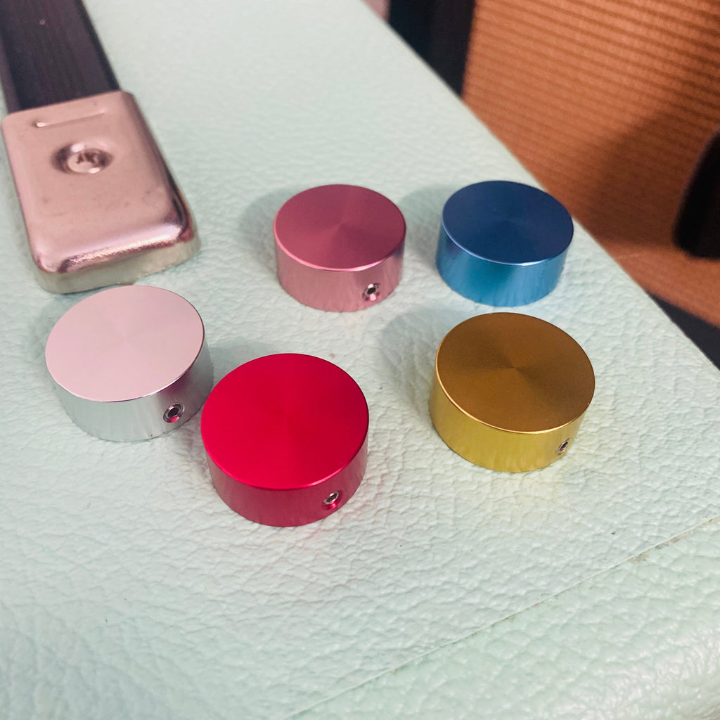 Five round metallic footswitch knob toppers in silver, pink, blue, red, and gold colors arranged on a textured surface.