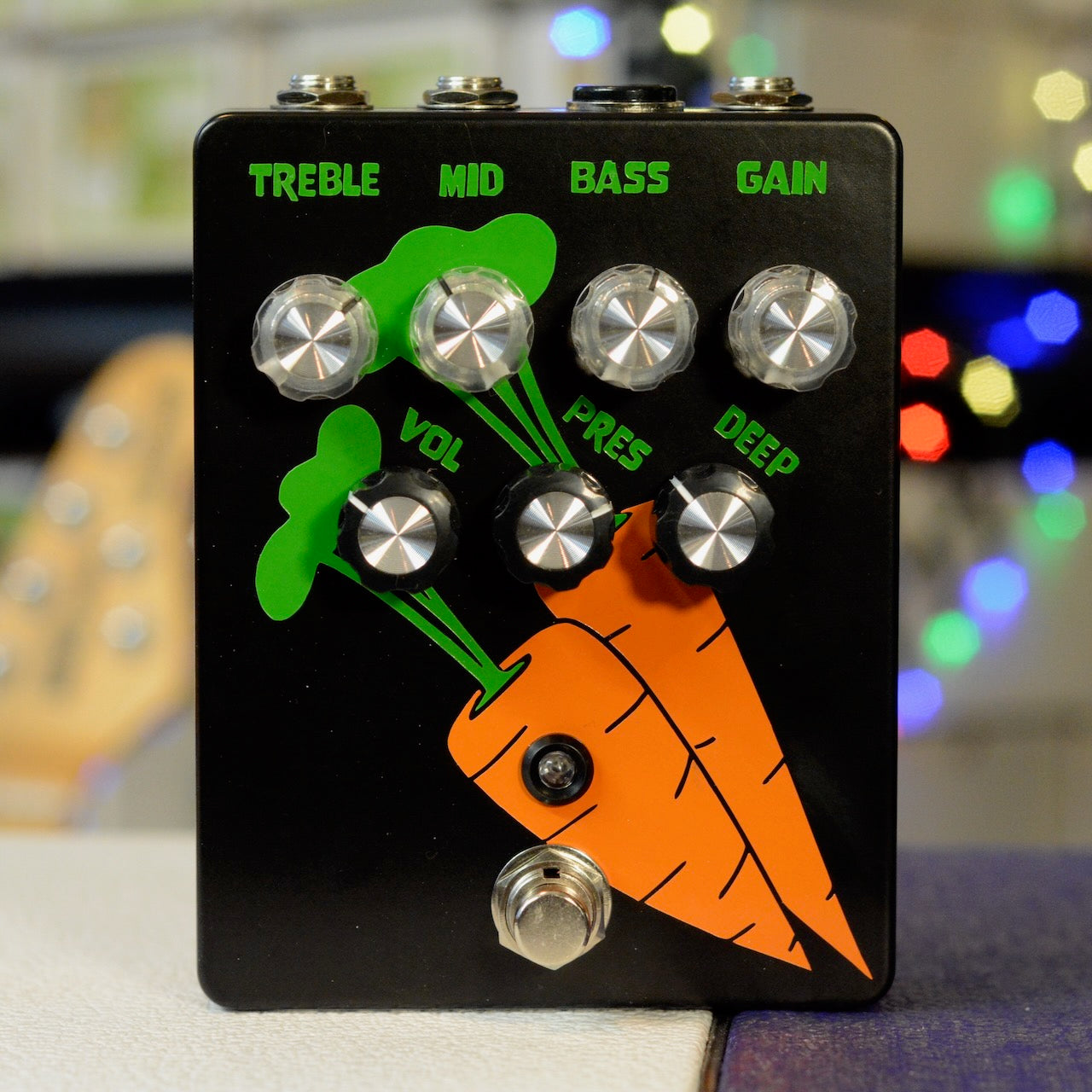 Black guitar pedal with green and orange carrot graphics and seven control knobs labeled with audio settings.