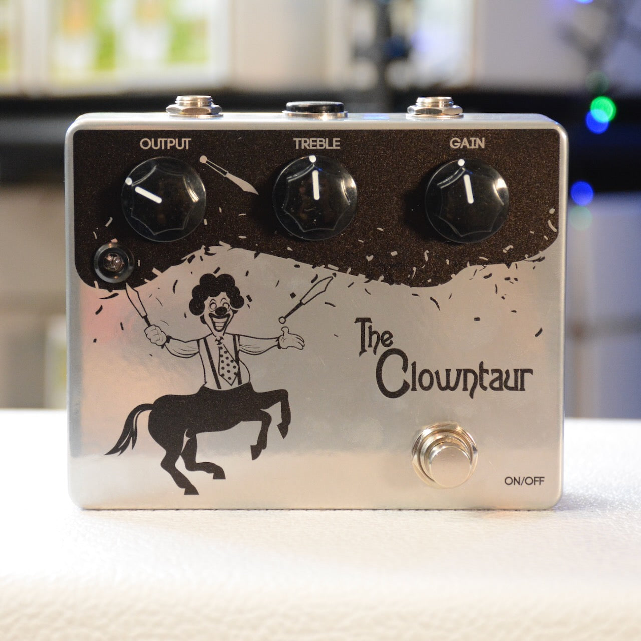 Silver guitar pedal with three black knobs labeled Output, Treble, Gain, a footswitch, and a centaur clown graphic.