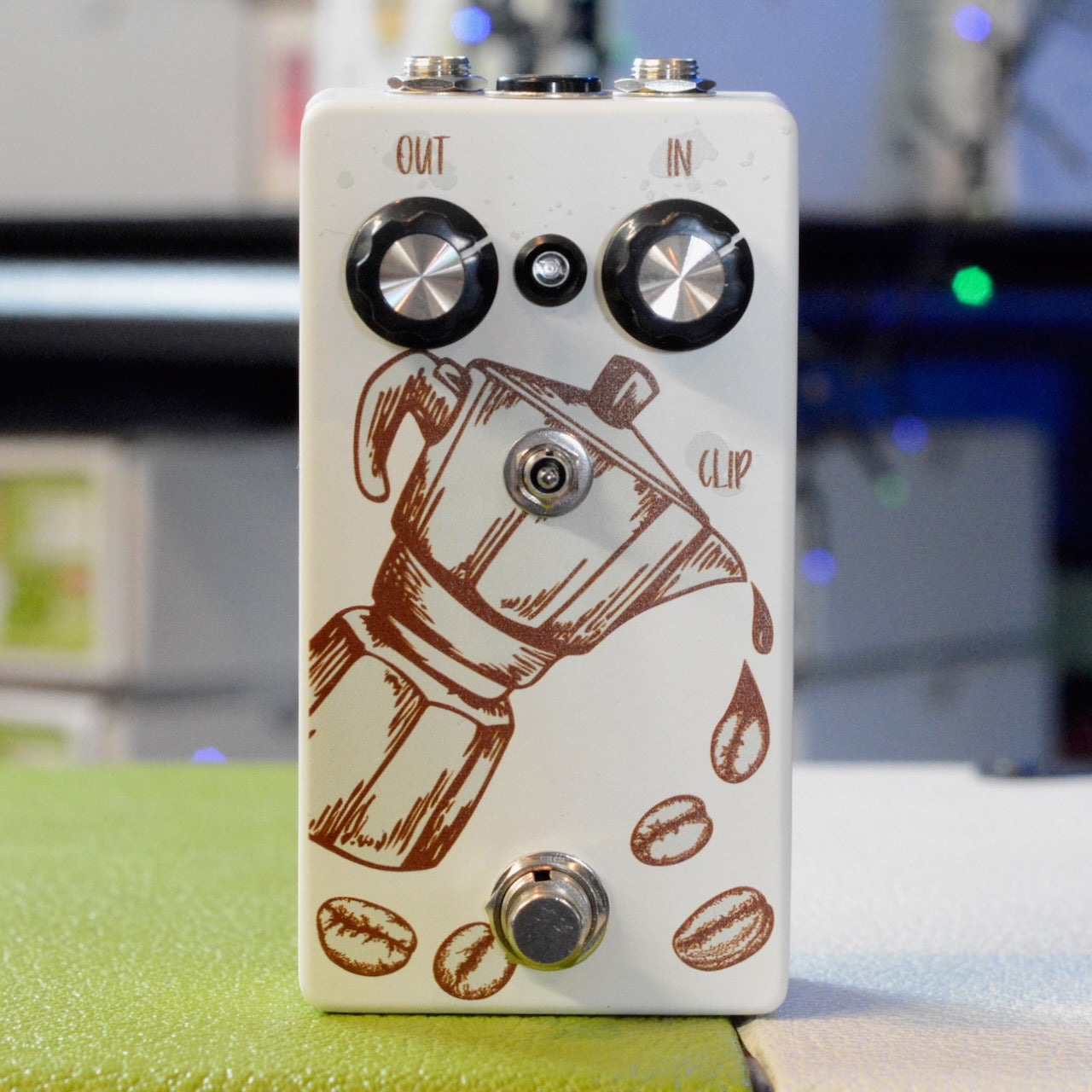 White guitar pedal with coffee pot graphic and coffee beans, two knobs labeled IN and OUT, a toggle switch, and a footswitch.