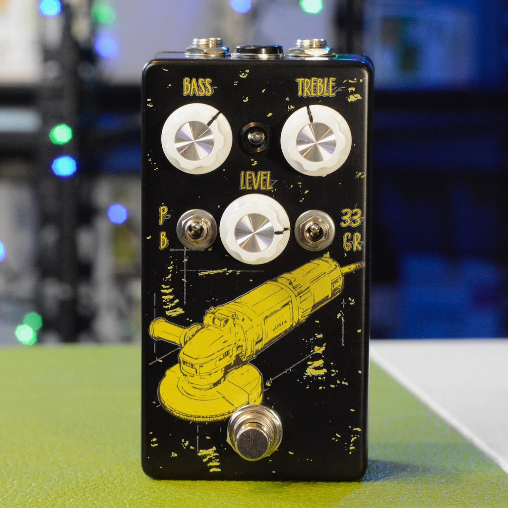 Black guitar pedal with three white knobs, two toggle switches, and footswitch featuring yellow grinder graphic.