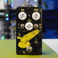 Black guitar pedal with three white knobs, two toggle switches, and footswitch featuring yellow grinder graphic.