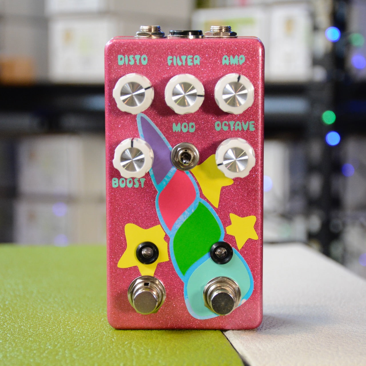 Front view of a pink guitar effects pedal with white knobs, toggle switch, footswitches, and colorful unicorn design.