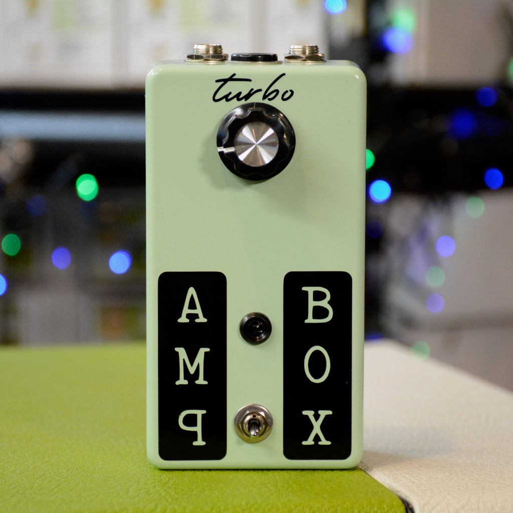 Light green pedal with one rotary knob, toggle switch, two quarter-inch jacks, and black labels reading AMP and BOX.