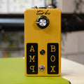 Gold-colored rectangular device with a rotary knob labeled turbo, toggle switch, and two 1/4 inch jacks labeled AMP and BOX.