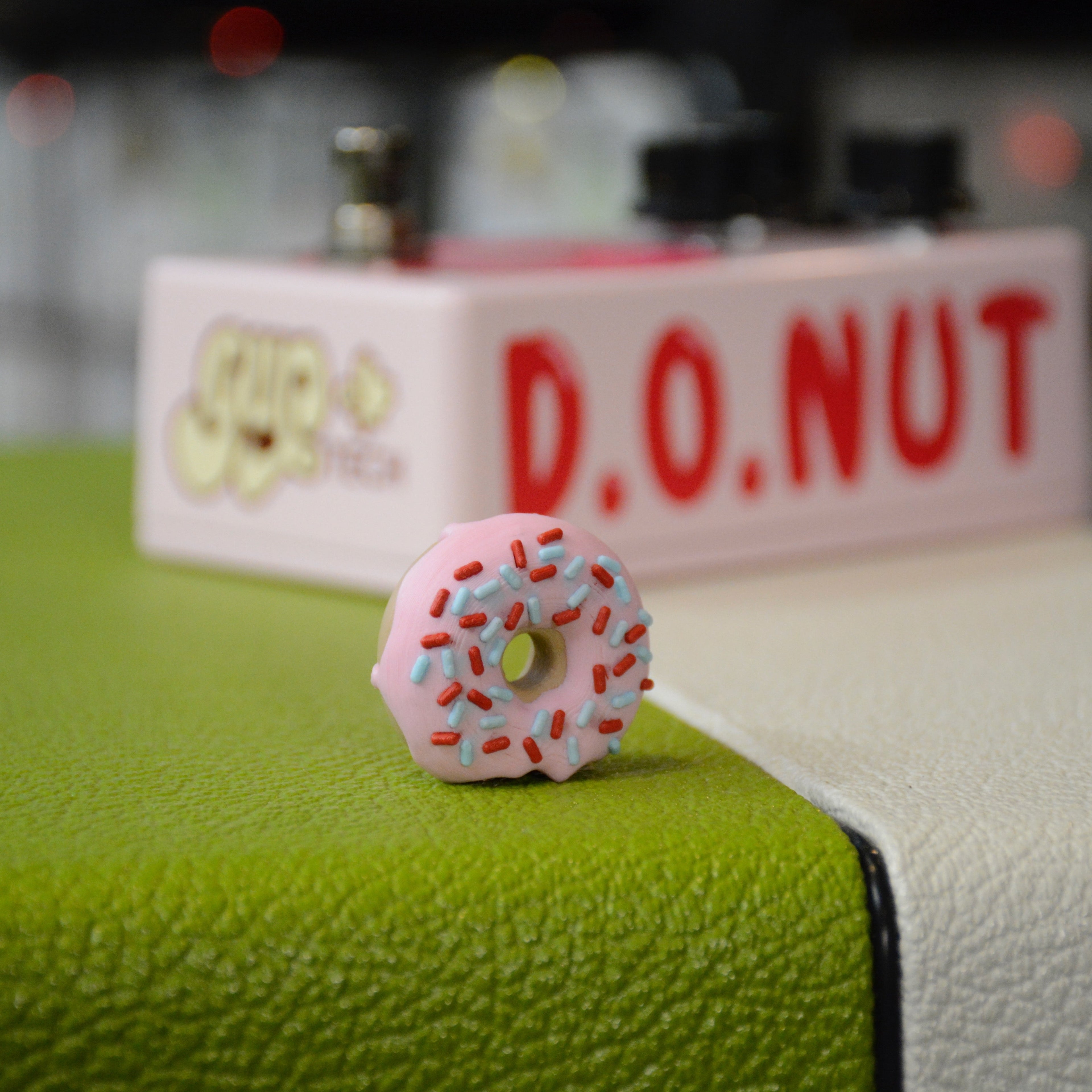 Small donut-shaped object with pink frosting and colorful sprinkles placed on a green and white surface.