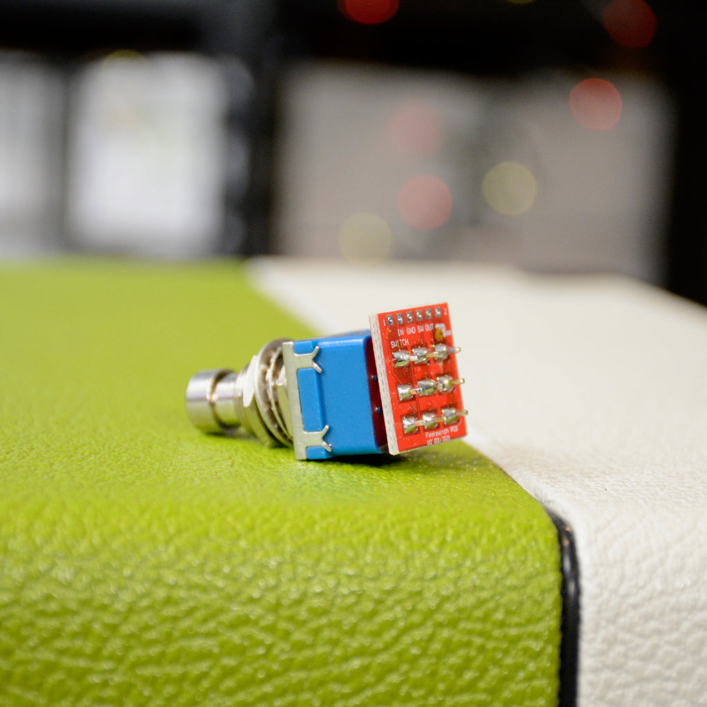 Close-up of a blue metal push-button switch with attached red circuit board and solder pins.