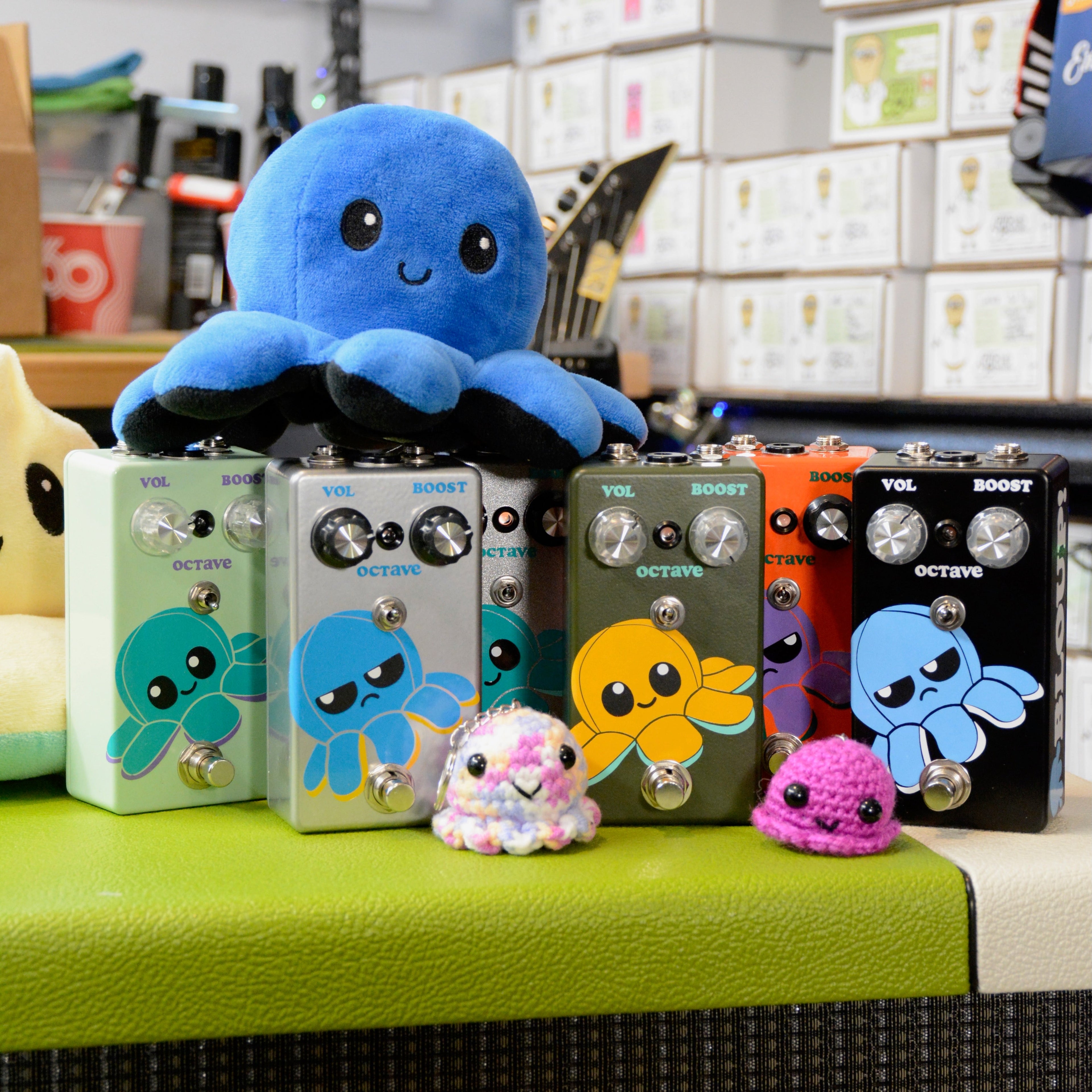 Six colorful guitar effects pedals with octopus graphics and control knobs arranged in a row with plush and crocheted octopus toys