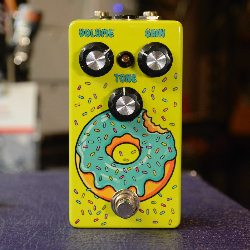A yellow guitar effects pedal with three black knobs labeled Volume, Gain, and Tone and a footswitch, featuring a cartoon donut graphic.
