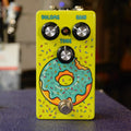 A yellow guitar effects pedal with three black knobs labeled Volume, Gain, and Tone and a footswitch, featuring a cartoon donut graphic.