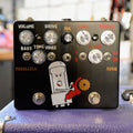 Black guitar effects pedal with multiple knobs, toggle switches, footswitches, and a cartoon tube character holding a saw.