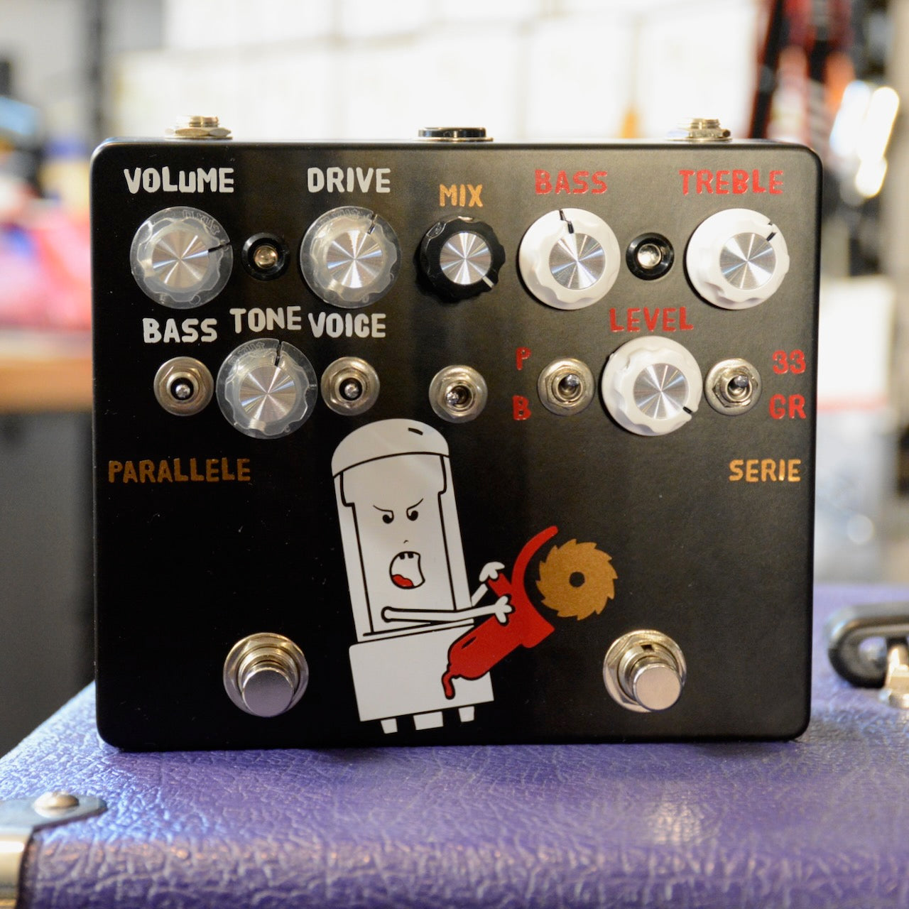 Black guitar effects pedal with multiple knobs, toggle switches, footswitches, and a cartoon tube character holding a saw.