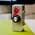 Silver rectangular device with black rotary knob and two audio jacks on the side with red graphics.
