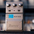 Gray Ibanez DE7 delay/echo guitar pedal with knobs, switches, and input/output jacks on a blurred background.