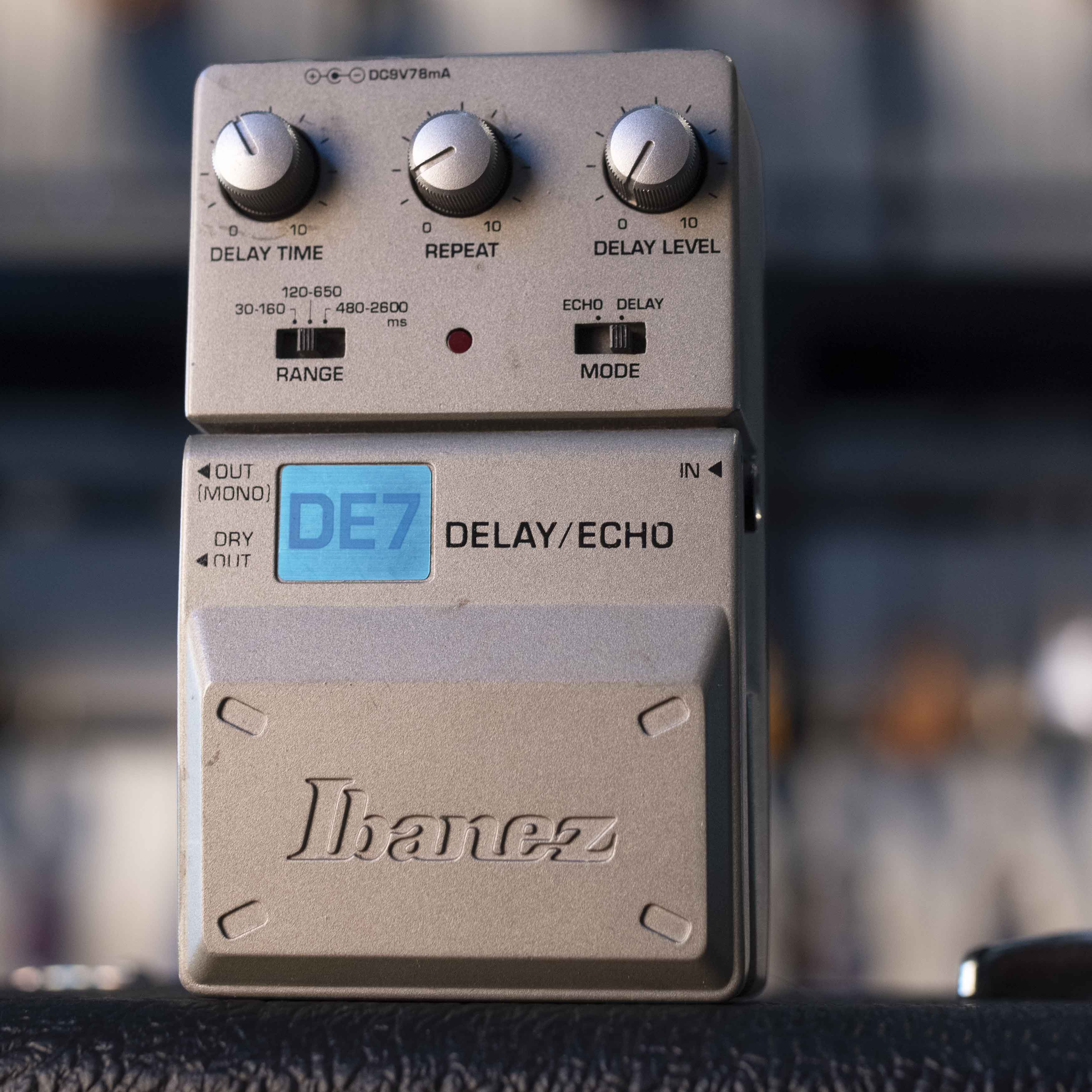 Gray Ibanez DE7 delay/echo guitar pedal with knobs, switches, and input/output jacks on a blurred background.
