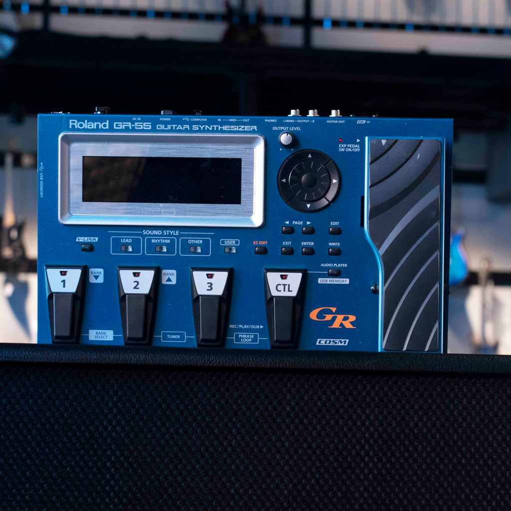 Blue Roland GR-55 guitar synthesizer with display, pedals, rotary dial, and multiple input/output ports.