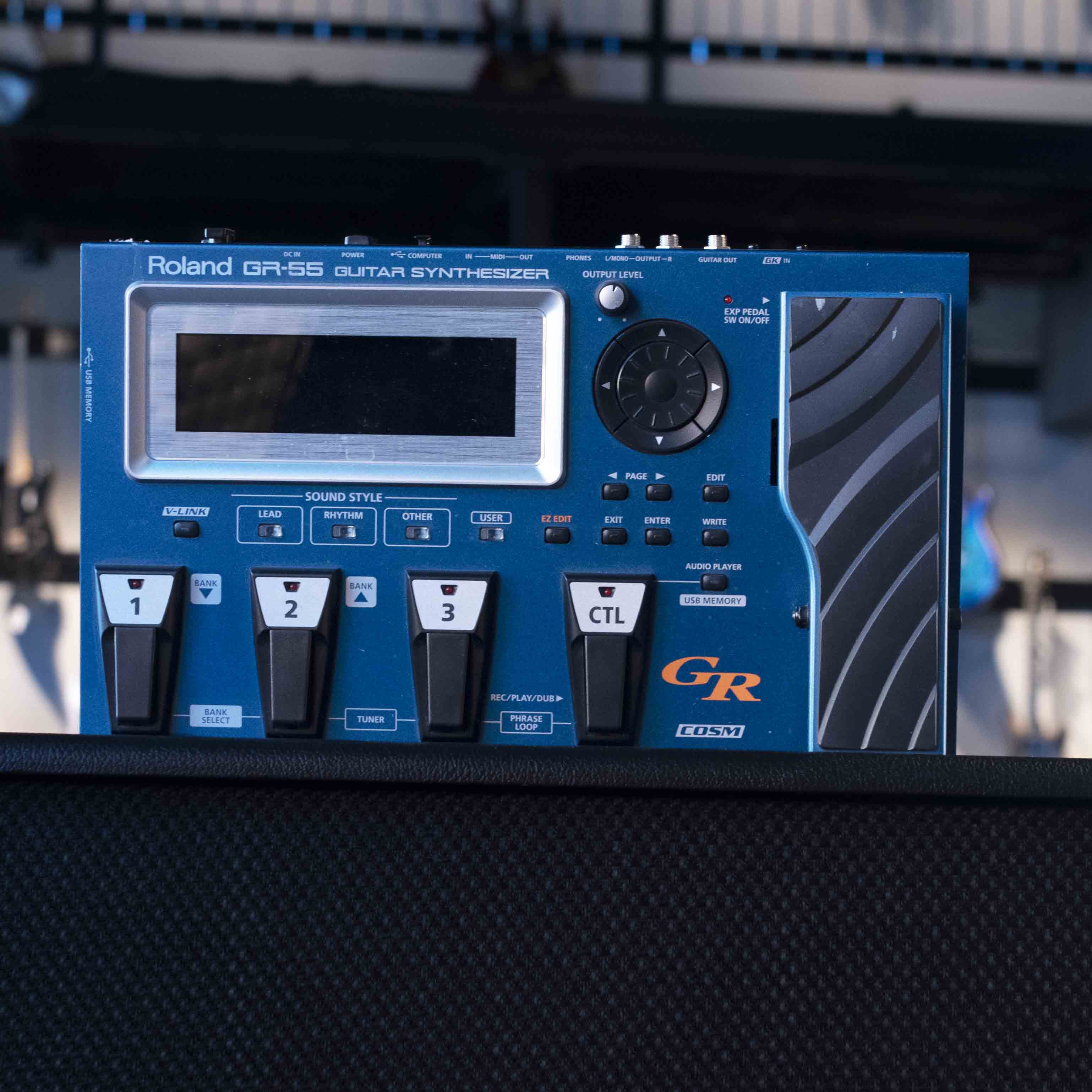 Blue Roland GR-55 guitar synthesizer with display, pedals, rotary dial, and multiple input/output ports.