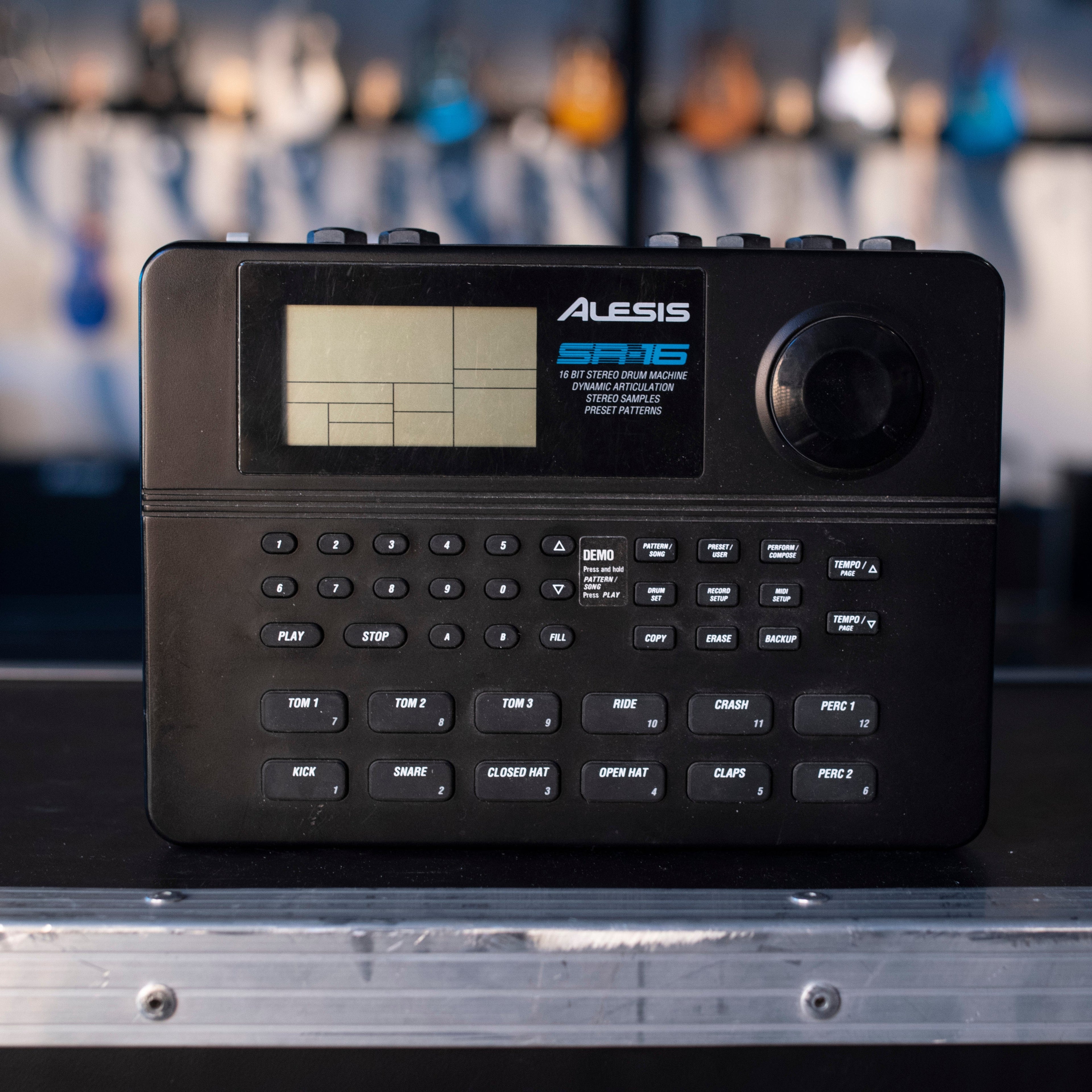 Black drum machine with buttons, a knob, and a screen showing Alesis SR16 branding and labels for drum sounds and controls.