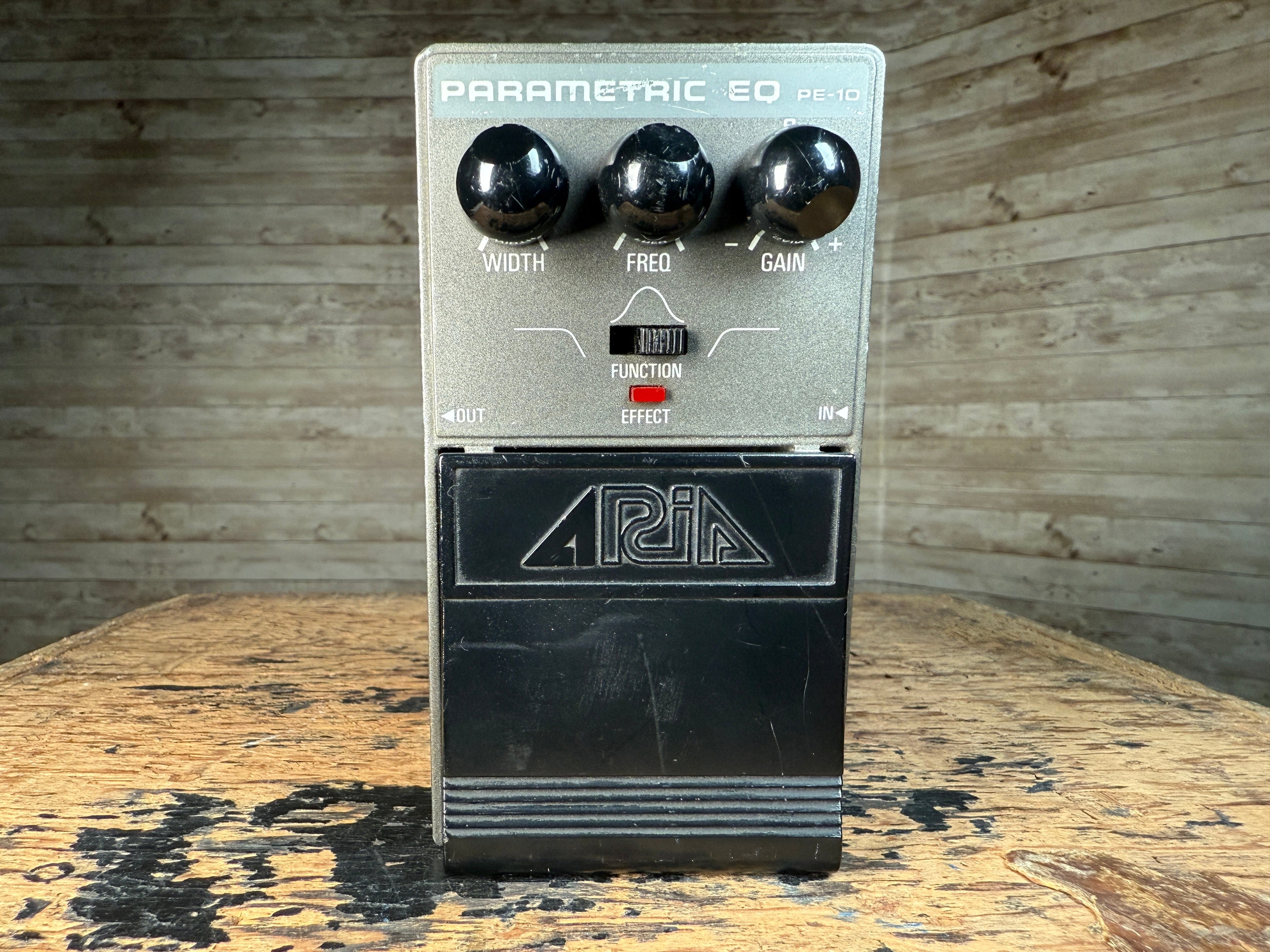Front view of a gray and black parametric equalizer pedal with three knobs and a switch on a wood surface.