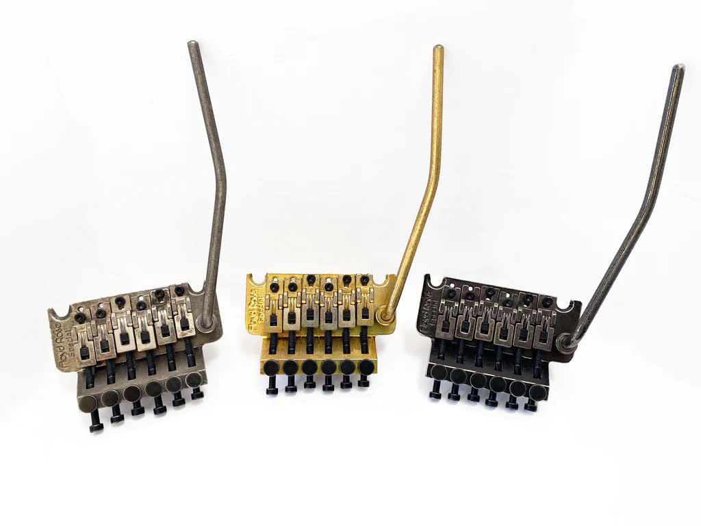 Three metal guitar tremolo bridge units with attached tremolo arms in silver, gold, and black finishes.