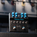 Black dual-sided overdrive pedal with blue knobs, toggle switches, and two footswitches on a textured surface.